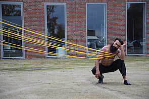 Ein Mann ist auf dem Rücken mit vielen gelben Schnüren verbunden und tanzt an ihnen auf dem Hof des Phantechnikums. 