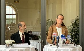 Dr. Michael Körner und Bettina Martin stehen an zwei runden Tischen mit weißen Tischdecken. Vor Bettina Martin stehen Mikrofone. 
