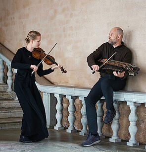 Ein Mann und eine Frau. Die Frau spielt stehend eine Barockvioline. Der Mann sitzt auf einem Treppengeländer und spielt eine Nyckelharpa.