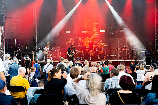 Eine Band spielt auf einer Bühne. Vor der Bühne stehen viele Zuschauer.