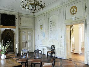 In einem Raum stehen vier dunkle Stühle und ein Tisch. An der Decke hängt ein Kronleuchter. Die Wände sind alt. An ihnen sind ein FDJ-Symbol und das Staatswappen der DDR zu sehen.