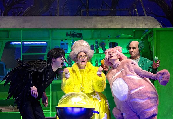 Vier Leute stehen auf einer Bühne. Sie tragen bunte Kostüme. Hinter ihnen steht ein grünes Regal. 