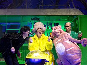 Vier Leute stehen auf einer Bühne. Sie tragen bunte Kostüme. Hinter ihnen steht ein grünes Regal.