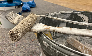 Auf einem leeren Farbeimer liegen ein benutzer Pinsel und eine Malerrolle.