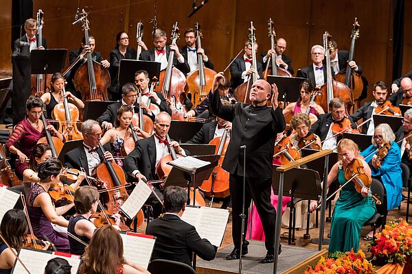 Jaap van Zweden steht dirigierend inmitten des New York Philharmonic Orchestra.