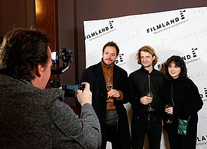 Zwei Männer und eine Frau stehen vor einer Interviewwand zum Filmkunstfest und werden fotografiert. 
