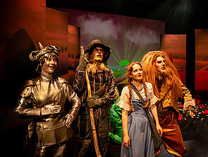 Das Foto zeigt vier verkleidete Menschen auf einer Bühne: einen eisernen Holzfäller, einen Strohmann, ein Mädchen, einen Löwen.