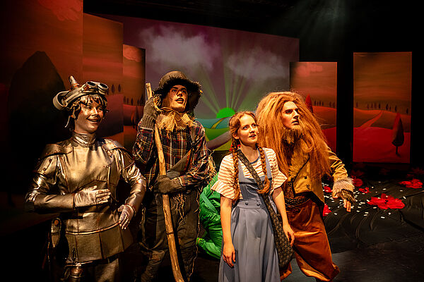 Das Foto zeigt vier verkleidete Menschen auf einer Bühne: einen eisernen Holzfäller, einen Strohmann, ein Mädchen, einen Löwen.
