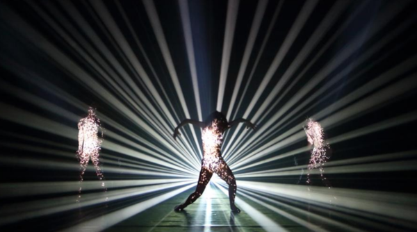 Drei Tänzer bewegen sich in leuchtenden Kostümen auf einer dunklen Bühne. Hinter ihnen bildet ein Scheinwerfer weiße Strahlen.