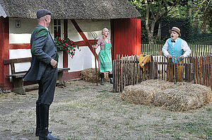 Ein Bauernhaus. Davor ist ein Garten. Im Garten stehen zwei Männer und eine Frau.