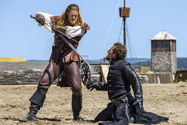 Ein Mann steht mit einem Schwert in der Hand am Strand. Ein Mann kniet vor ihm im Sand.