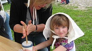 Ein Kind sitzt an einem Tisch. Es trägt eine historische Kopfhaube und hält einen Becher in der Hand. Eine Frau reibt zwischen beiden Händen einen Quirl im Becher.