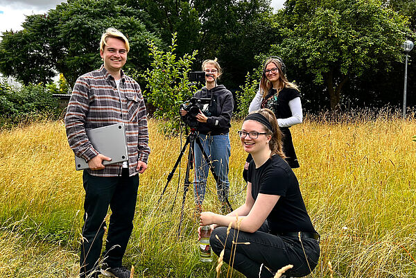 Zwei Männer und zwei Frauen stehen oder hocken auf einer Wiese. Einer der Männer hat einen Laptop unterm Arm, der andere steht hinter eine Kamera.