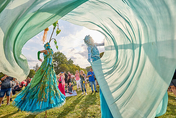 Zwei Künstler auf Stelzen. Sie tragen bunte Gewänder. Sie stehen auf einer Wiese.