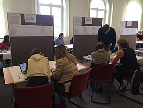 Männer und Frauen sitzen an Tischen, blicken auf Laptops und Zettel. Vor ihnen stehen drei Rolltafeln, an denen ebenfalls Zettel hängen.