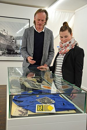 Ein Mann und eine Frau stehen an einer Vitrine mit Exponaten aus DDR-Zeiten. Darunter befinden sich ein FDJ-Hemd und alte Fotos.