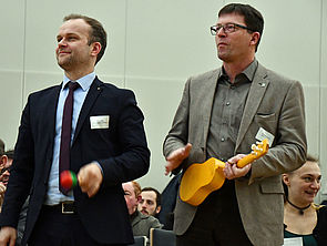 Zwei Teilnehmer der Konferenz stehen im Raum und haben eine Rassel und eine Ukulele in der Hand.