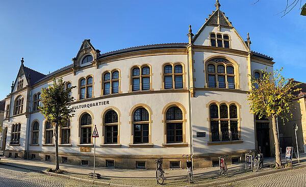 Außenfassade vom Kulturquartier. Das Gebäude ist zwei Stockwerke hoch. In unregelmäßigen Abständen kommt ein spitzer Giebel dazu. Auf beiden Etagen reihen sich Fenster an Fenster. Auf der hellen Fassade steht in dunkler Schrift "Kulturquartier".