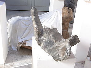 Eine Skulptur ist mit einem weißen Tuch verhüllt. Eine andere liegt auf einem weißen Sockel.