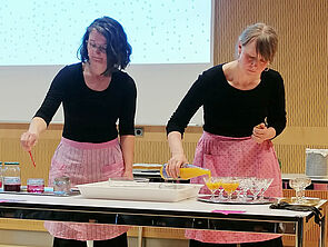 Zwei Frauen mit Schürzen an einem Tisch. Sie bereiten etwas zu.
