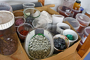 Ein Karton voller Becher. Darin befinden sich verschieden farbige Granulate.