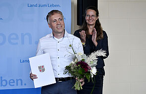 Thomas Schmidt hält eine Urkunde und Blumen in den Händen. Hinter ihm steht Bettina Martin und applaudiert.