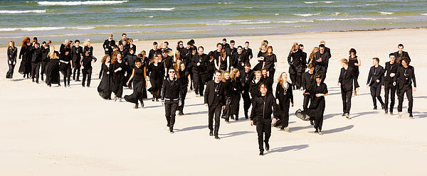 Die Mitglieder des Baltic Sea Philharmonic stehen schwarz gekleidet am Stand. Hinter ihnen schlägt die Ostsee Wellen.