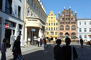 Menschen gehen über den Marktplatz von Stralsund. Am Rand stehen Giebelhäuser in weiß, gelb und braun.