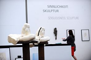 Auf zwei braunen Sockeln stehen zwei weiße Skulpturen. Im Hintergrund fotografiert eine Frau die Ausstellung.