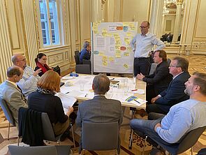 Ein Tisch voller Zettel. Darum verteilen sich neun Frauen und Männer. Ein Mann steht am Tisch neben einer Magnetwand, an der ebenfalls viele Zettel hängen.