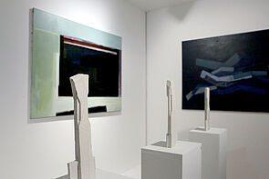 Zwei dunkle Bilder an der Wand. Davor sind drei Skulpturen zu sehen.