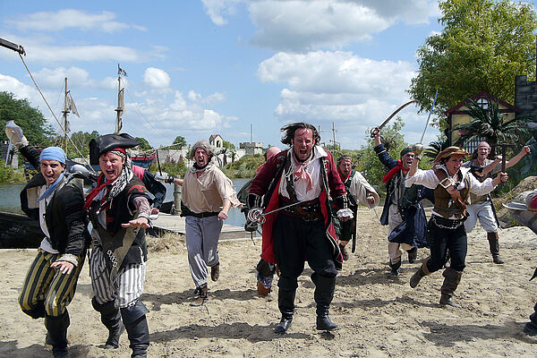 Piraten stehen im Strandsand. Im Hintergrund Wasser und Bäume.