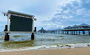 In der Ostsee, auf zwei Pflöcken, steht eine große Videoleinwand. Im Vordergrund ist Sand. Im Hintergrund steht die Seebrücke von Heringsdorf.