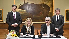 Manuela Schwesig und Alexander Otto sitzen an einem Tisch. Vor ihnen liegen Dokumente. Beide haben einen Stift in der Hand. Neben Manuela Schwesig steht Reinhard Meyer. Neben Alexander Otto steht Rico Badenschier. Im Hintergrund hängt eine großes Gemälde von Nashorn Clara an der Wand.