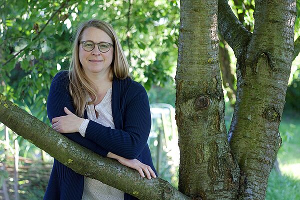 Porträt einer Frau. Sie steht an einem Baum und lehnt auf einem Ast.