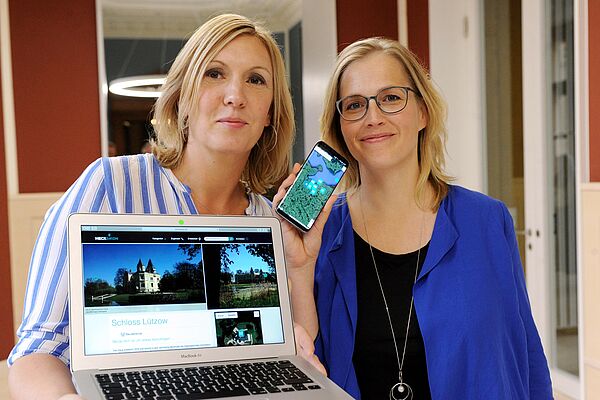 Zwei Frauen stehen nebeneinander. Eine von ihnen hält einen aufgeklappten Laptop in der Hand, die andere ein Handy. 