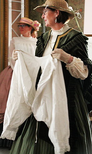 Eine Frau in historischer Kleidung hält eine Beinhose in der Hand. 