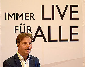 Ein Porträt vonMark Rohde. Hinter ihm, auf einem Aufsteller, steht: "Immer live für alle."