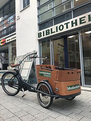 Vor dem Eingang der Bibliothek steht ein Lastenfahrrad mit der Aufschrift "Medienbote". 