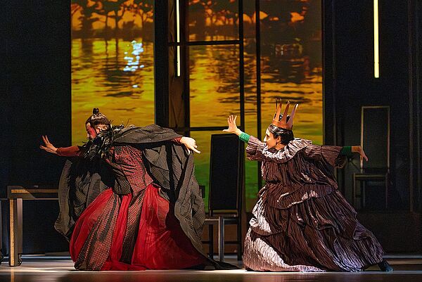 Ein Mann und eine Frau. Sie stehen auf einer Bühne. Sie tragen opulente Kleider. 