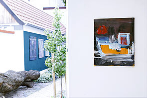 Rechts hängt ein Bild aus der Ausstellung. Links ist ein blaues Haus zu erkennen.
