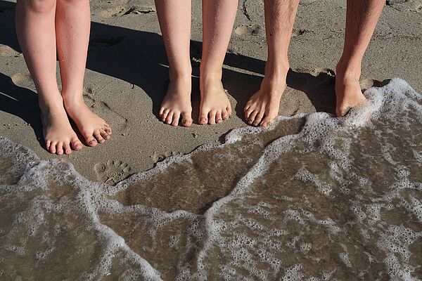 Sechs nackte Füße stehen im Sand. Wasser kommt und umspült sie.