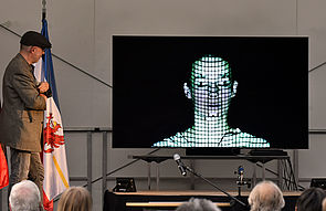 Dr. Marcus Doering steht links auf einer Bühne. Rechts neben ihm ein Flachbildschirm mit dem weißen Porträt eines Mannes. Vor der Bühne sitzen Zuschauer und Zuschauerinnen.