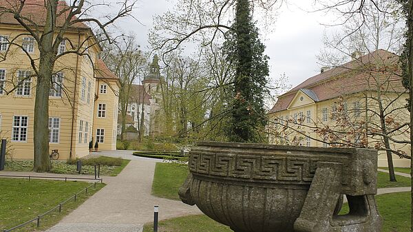 Ein Park mit Bäumen. Im Vordergrund steht ein Brunnen. Im Hintergrund verteilen sich drei helle Gebäude.