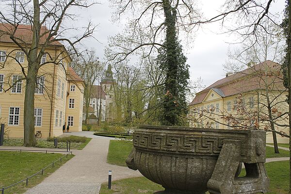 Ein Park mit Bäumen. Im Vordergrund steht ein Brunnen. Im Hintergrund verteilen sich drei helle Gebäude.