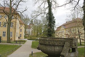 Ein Park mit Bäumen. Im Vordergrund steht ein Brunnen. Im Hintergrund verteilen sich drei helle Gebäude.