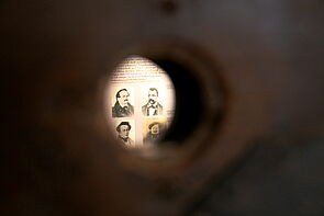 Ein Guckloch. Dahinter: der Ausschnitt einer Infotafel.