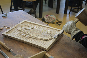 Eine Hand hält ein Schnitzwerkzeug und bearbeitet ein Holzbrett. Auf dem Brett zeichnet sich das Relief eines Seepferdchens ab.