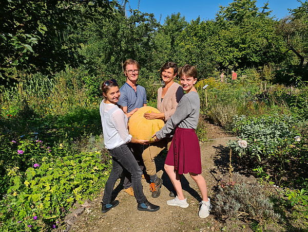 Vier Jugendliche stehen in einem Garten und halten gemeinsam einen großen, gelben Kürbis fest.