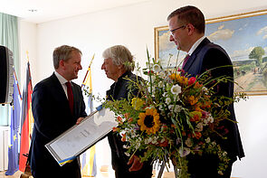 Zwei Männer überreichen eine Urkunde an einen dritten.